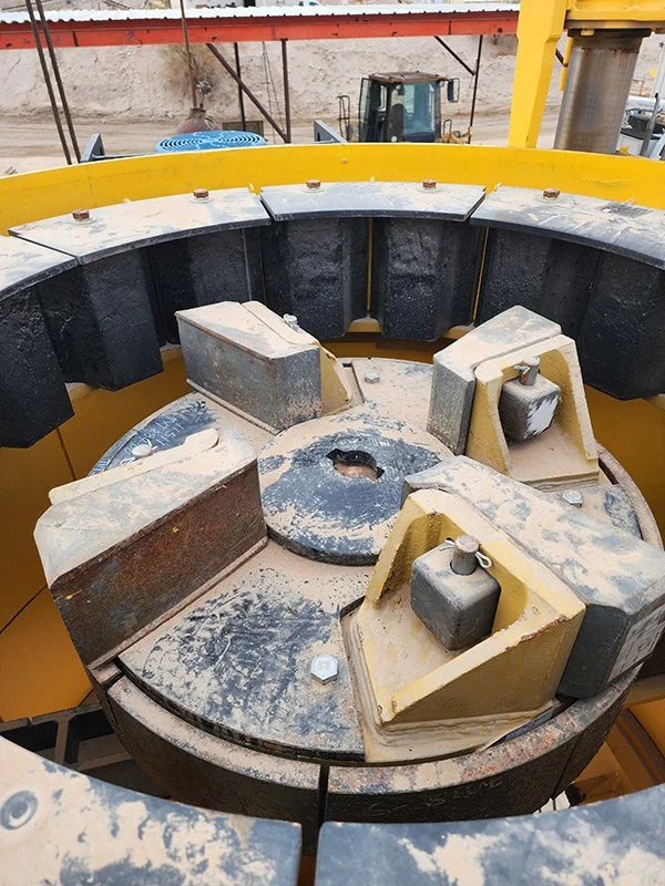 Close-up of a large industrial machine part with metal blocks and rubber or plastic inserts, dust and dirt, at a construction site with heavy machinery and construction materials.