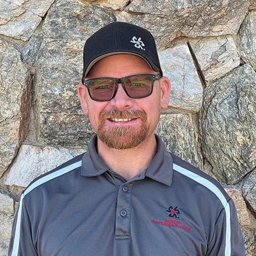 A man with a beard, wearing a black cap with a logo, sunglasses, and a gray collared shirt with a logo on it, standing in front of a stone wall.