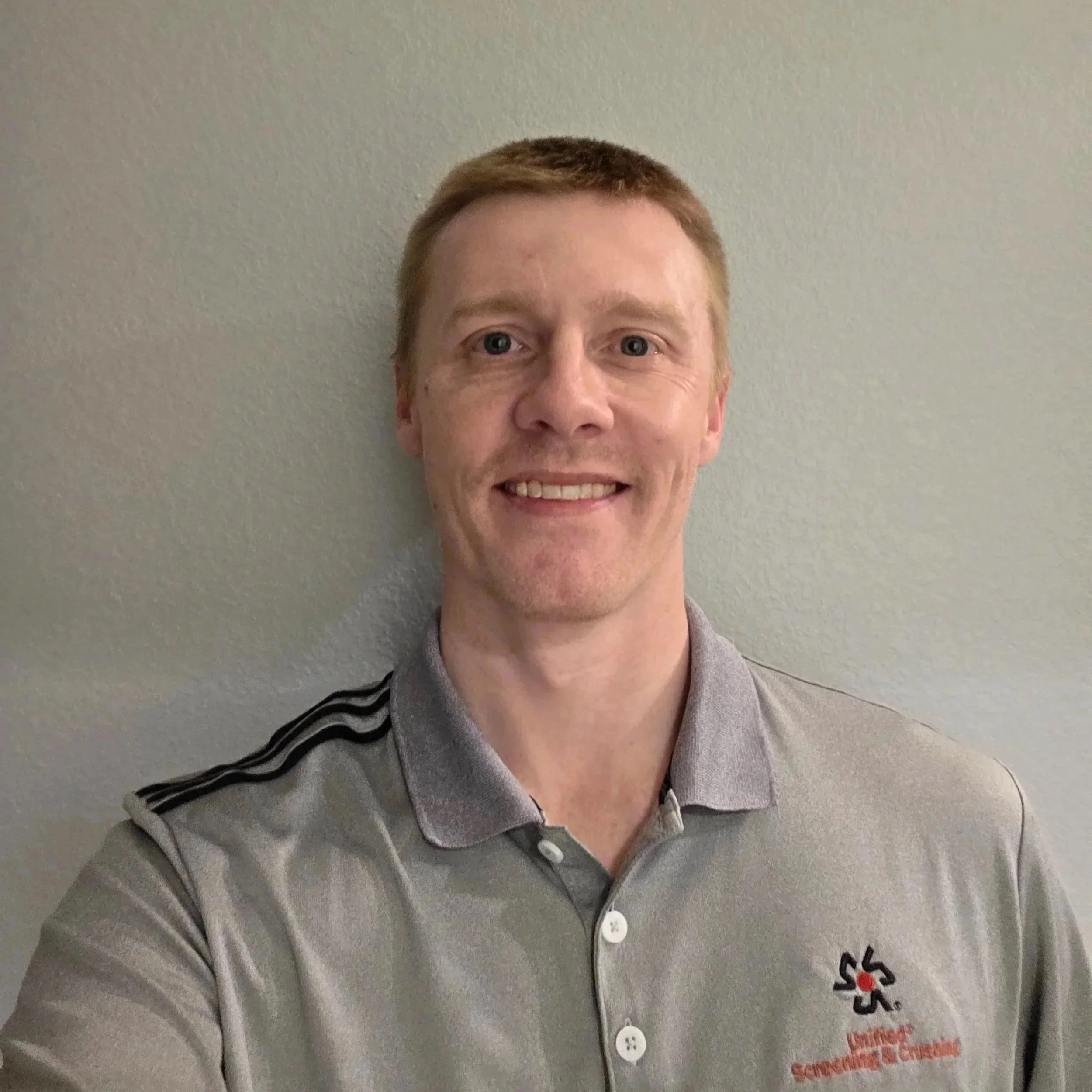 Close-up of a person's face and upper body, wearing a gray chef jacket with black and red embroidered words, standing against a plain light-colored wall.
