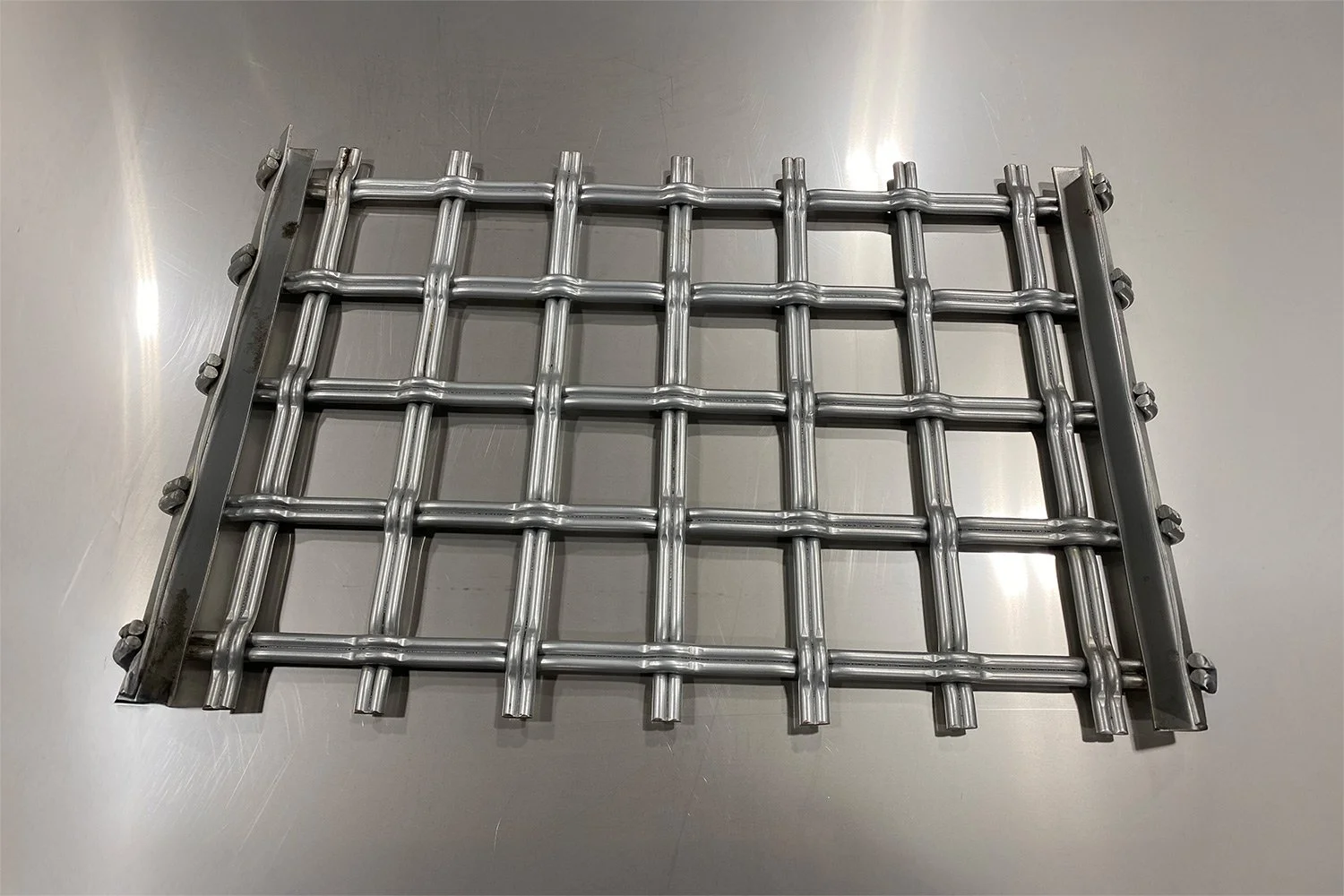 A metal grid with a rectangular shape, consisting of horizontal and vertical bars, mounted on a flat metallic surface.