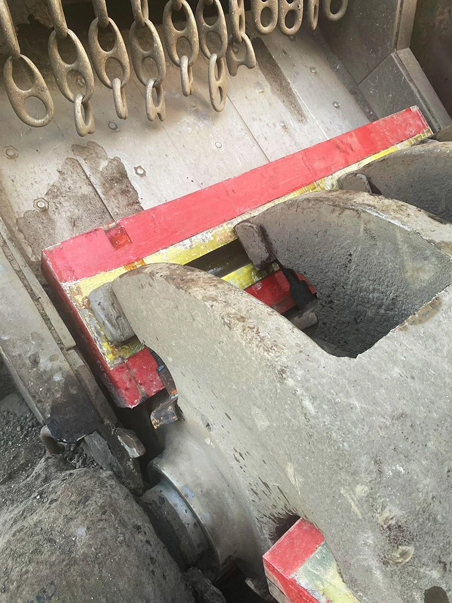 Close-up view of a large industrial machine component with red and yellow painted edges, showing wear and grime, with metal hooks hanging above.