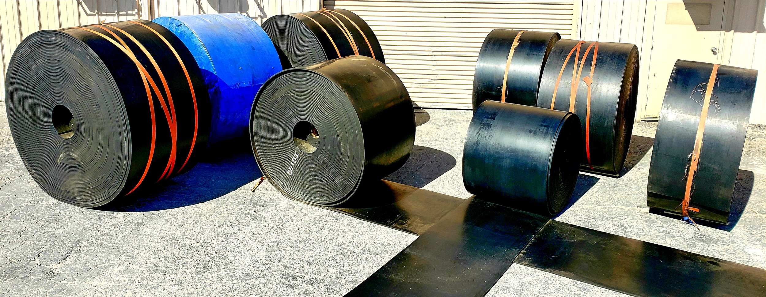 Several large rolls of black and blue rubber or plastic material stacked outdoors on a concrete surface near a metal building.