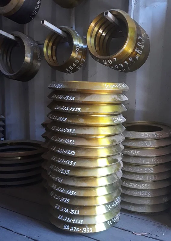 Stacked large brass or gold-colored mechanical parts with serial numbers on a shelf in an industrial setting.