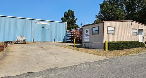 A small beige building with windows and a sign next to a large blue warehouse with a basketball hoop and a trash container outside. There are trees and a clear blue sky in the background.
