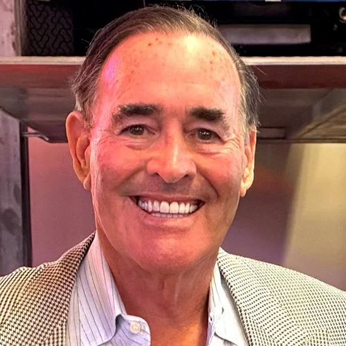 A smiling middle-aged man with gray hair, wearing a light-colored blazer and striped dress shirt, standing indoors.