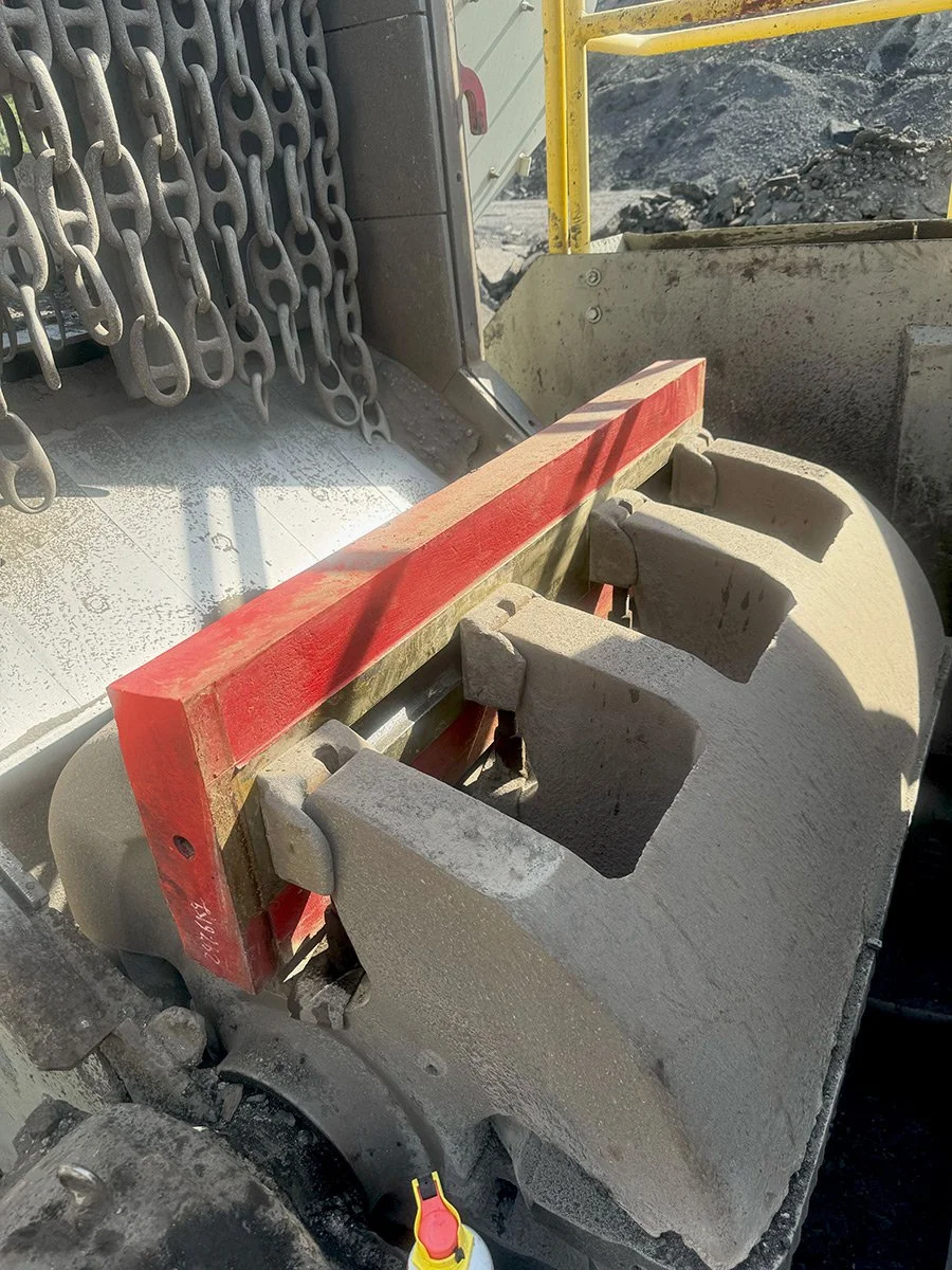 Close-up view of a large industrial concrete paving or cement cutting machine with metallic chains on the left side, and empty mold cavities for concrete blocks on the right, in an outdoor construction site with dirt and gravel.