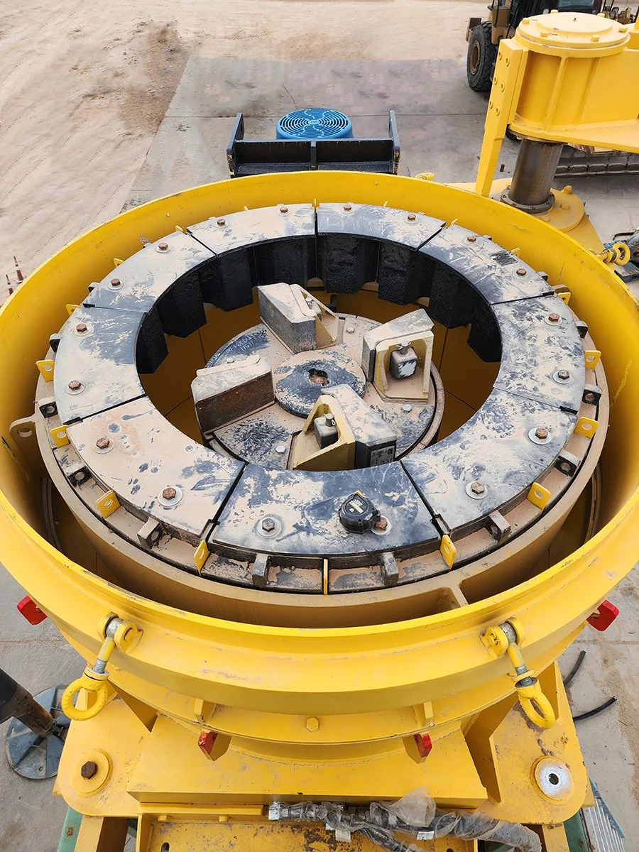 Close-up of a large, yellow construction machine component with a circular, segmented metal ring and black cushioning pads. The background shows part of a construction site with dirt, a blue fan, and other machinery.