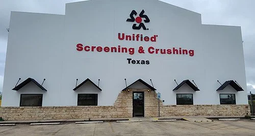 The exterior of a building with a sign reading "Unified Screening & Crushing Texas" and six small roofed windows.