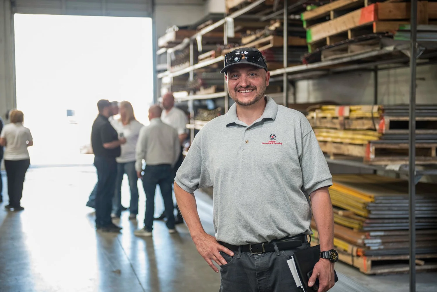 Smiling man wearing a gray polo shirt and sunglasses on his hat stands inside a warehouse with other people in the background, alongside shelves of wood and metal materials.