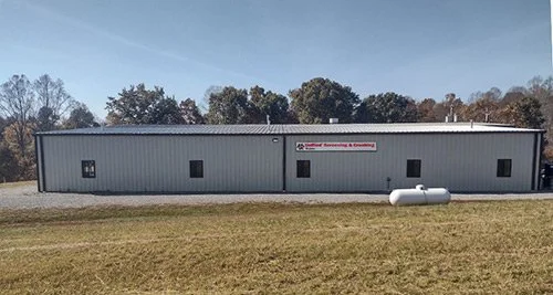 A long, rectangular metal building with a sign on the side, situated on a grassy area under a clear sky, with trees in the background.