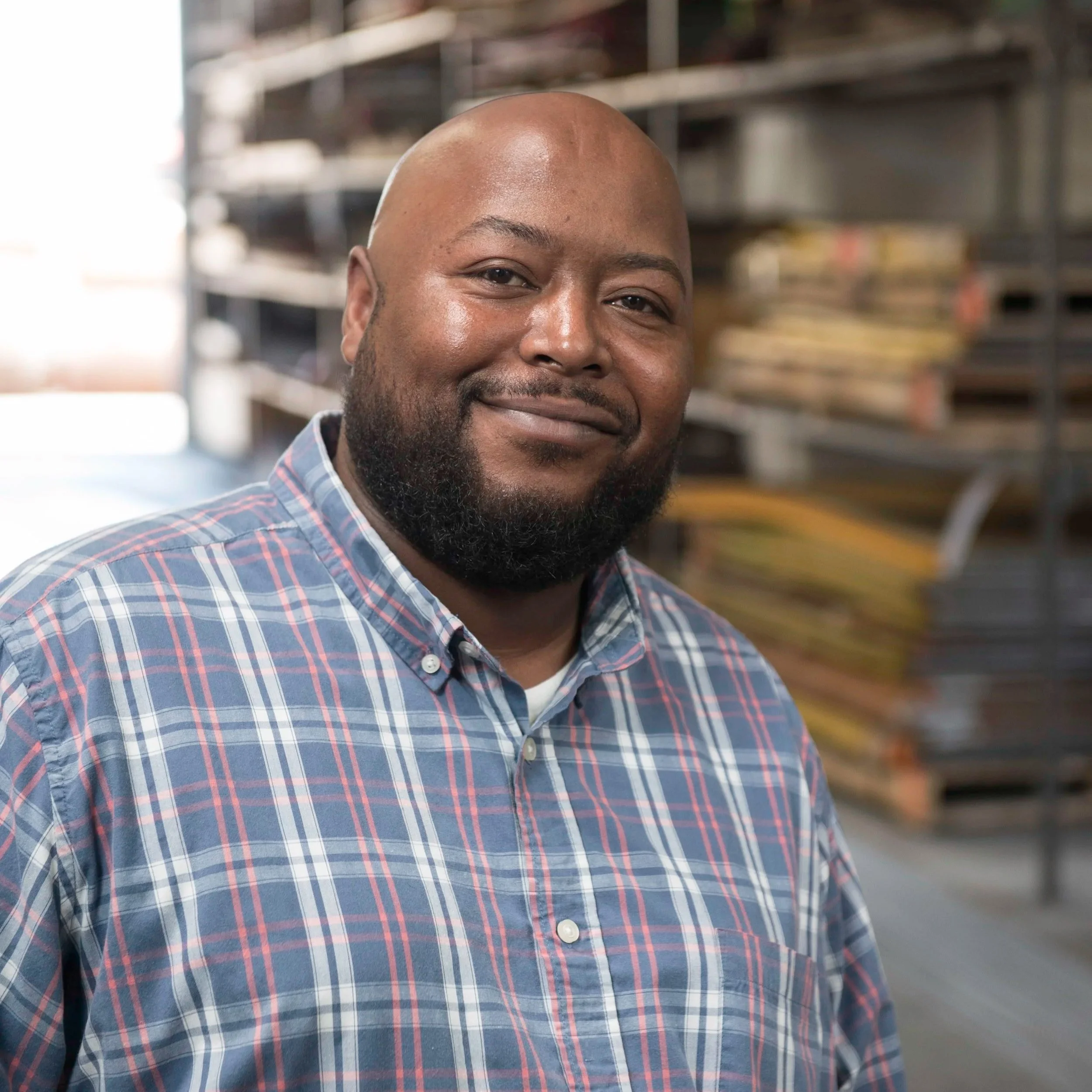 Close-up of a smiling African American man with a shaved head and beard, wearing a plaid shirt, standing in front of a blurred background of shelves in a store or warehouse.