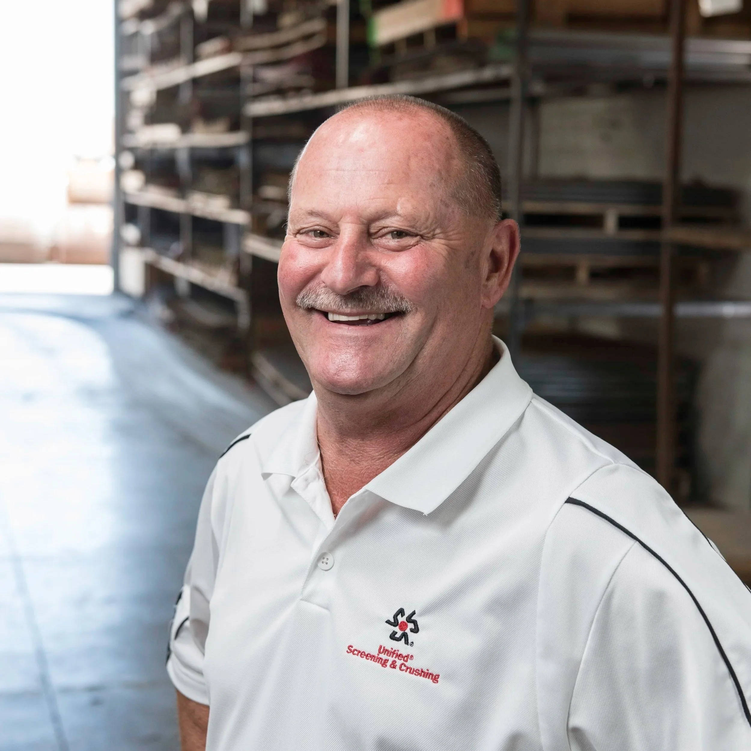 A smiling man with a bald head and mustache wearing a white shirt with black accents, standing in front of a shelving unit with multiple wooden pallets in an industrial or warehouse setting.