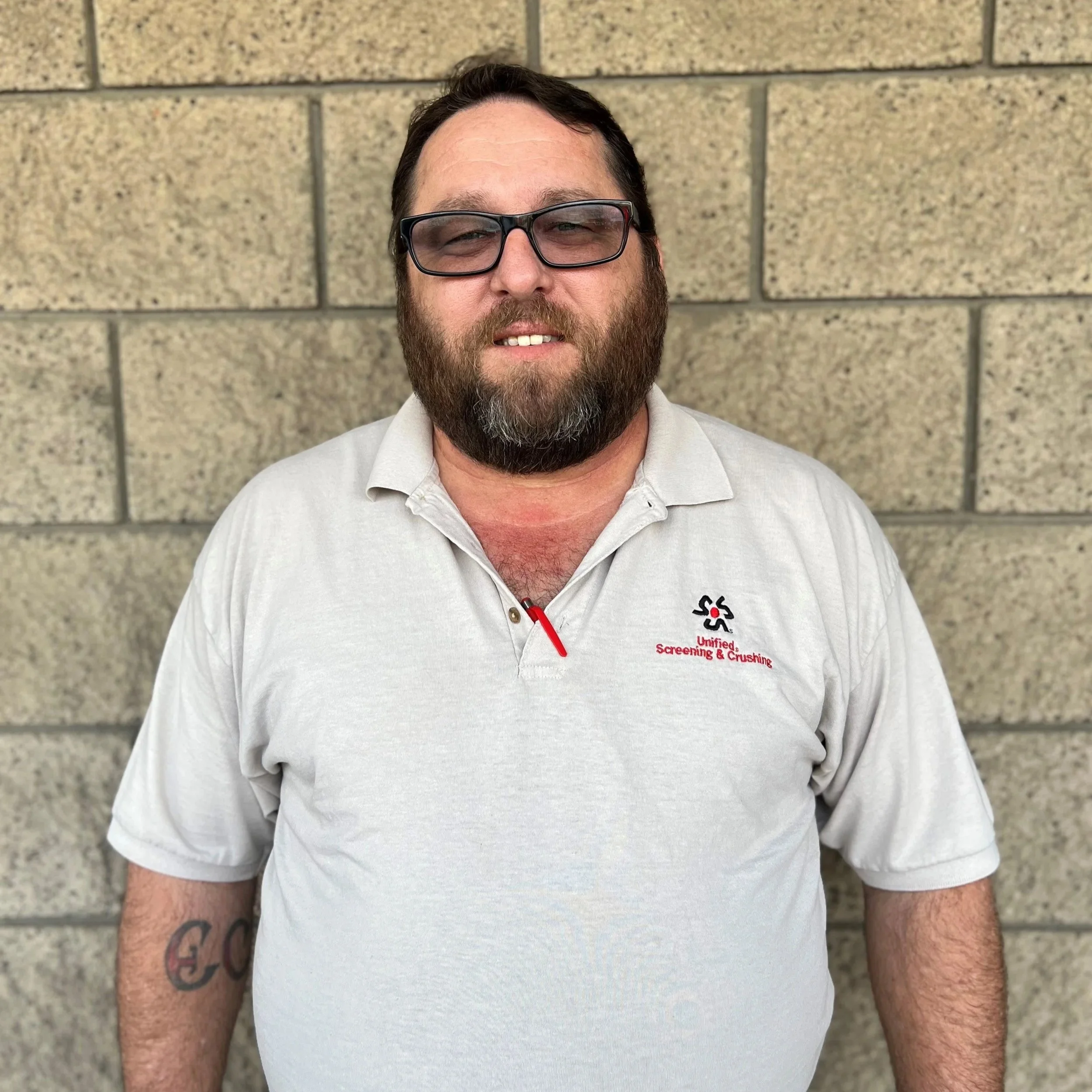 A man with a beard and glasses wearing a light-colored polo shirt, standing against a brick wall.