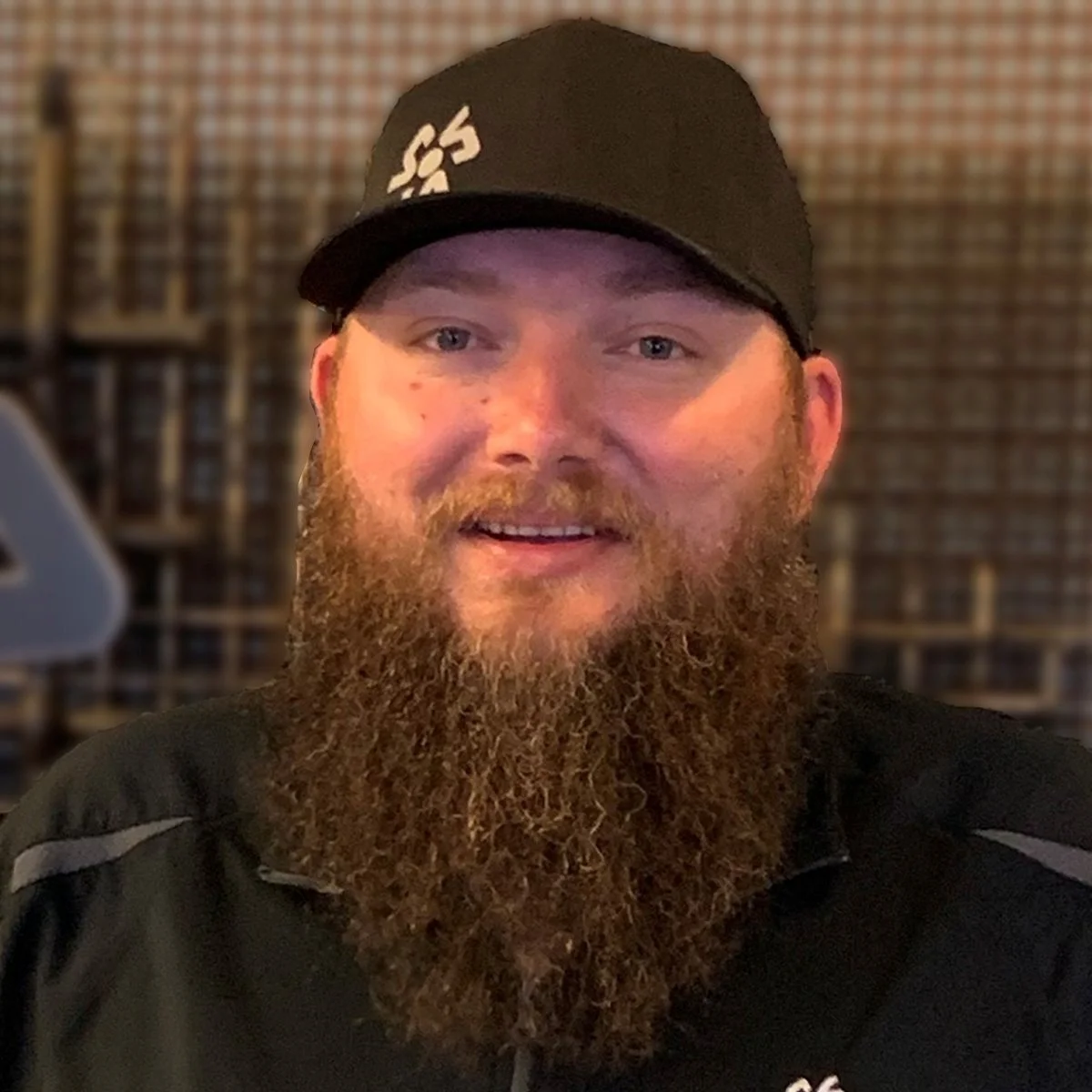 A man with a full beard wearing a black cap and black jacket, smiling indoors with a blurred background.