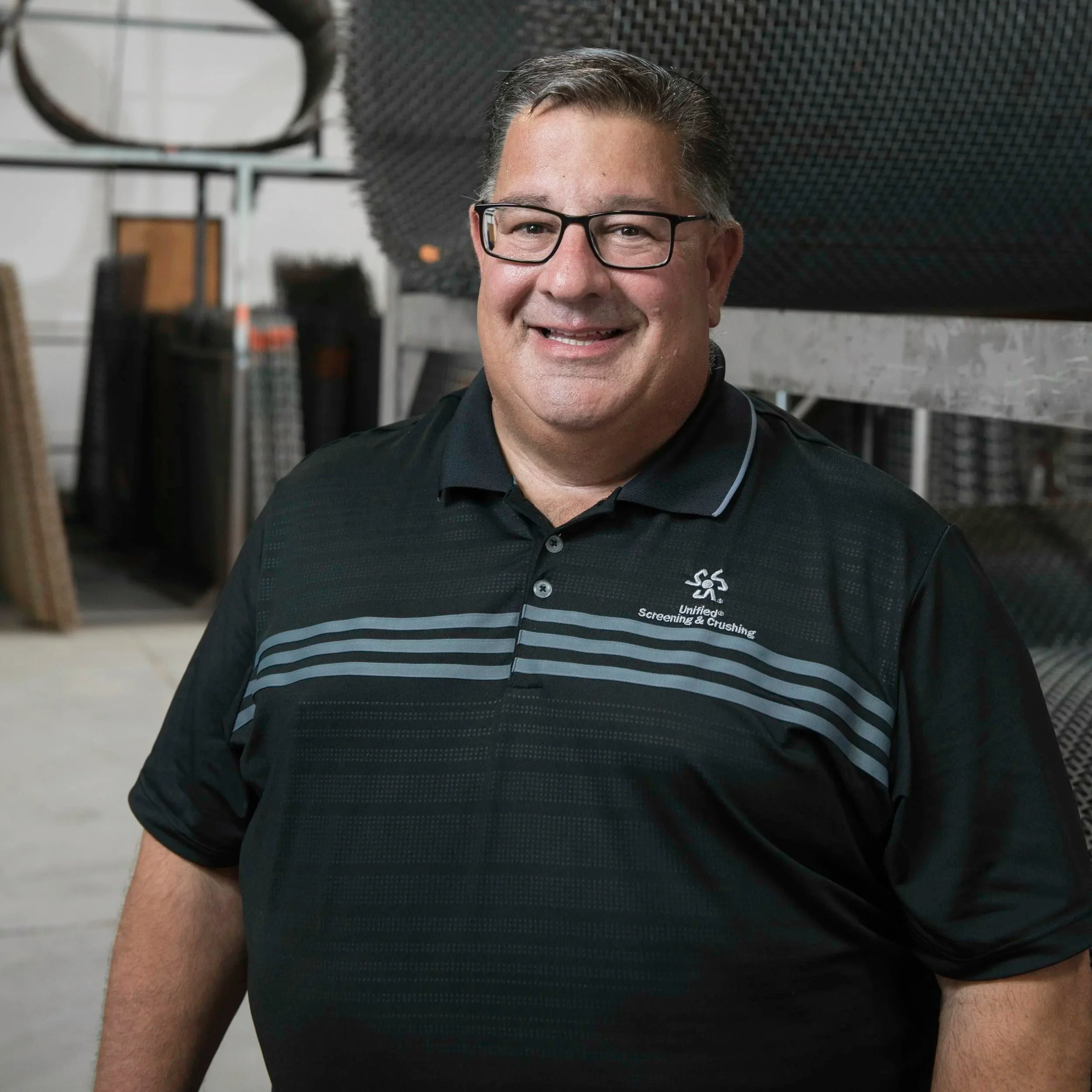 Close-up of a smiling man with glasses wearing a black shirt with a logo and company name, standing in an indoor industrial or warehouse setting.