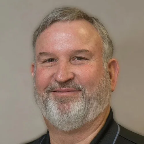 A smiling middle-aged man with a beard and short gray hair, wearing a dark collared shirt, against a plain background.