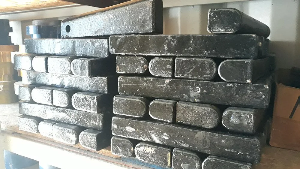 Stacked blocks of metal, possibly ingots, with a rough, worn surface, stored on a shelf in a workshop or industrial setting.