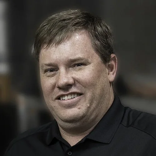 A man with light brown hair wearing a black polo shirt smiling at the camera.