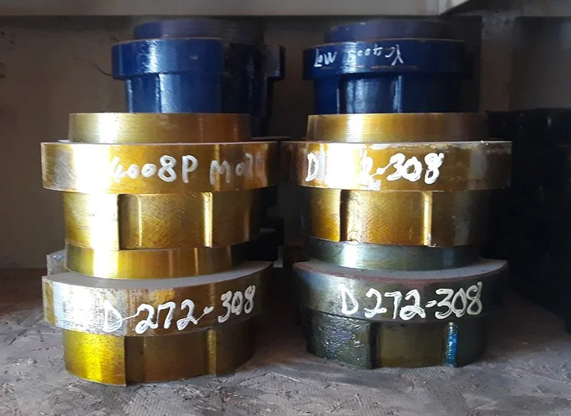 Stacked industrial metal machine parts with yellow and black coating, labeled with handwritten white markings, on a rough wooden surface.
