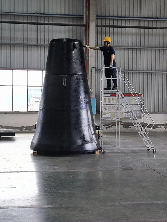 A person wearing a yellow safety helmet and gloves sprays a black cone-shaped object inside a spacious warehouse.