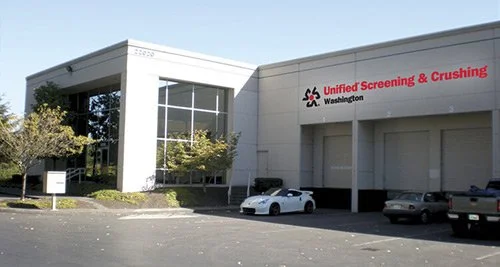 Exterior view of a building with a sign that reads 'Unified Screening & Crushing Washington' and several parked cars in front.