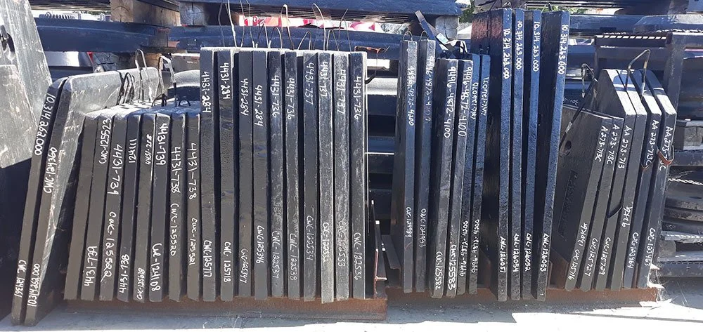 Multiple steel beams stacked outdoors on a truck bed, with white handwritten labels indicating different specifications.