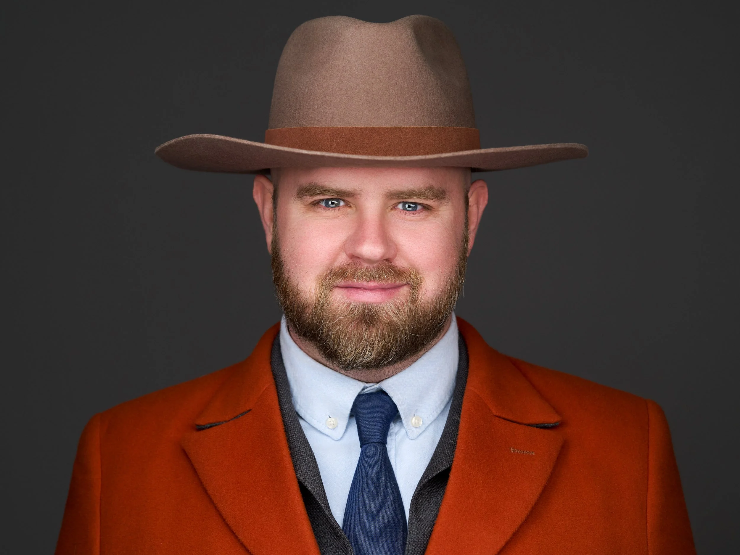 A man with a beard and blue eyes wearing a brown hat, a red coat, a white shirt, and a blue tie against a dark background.