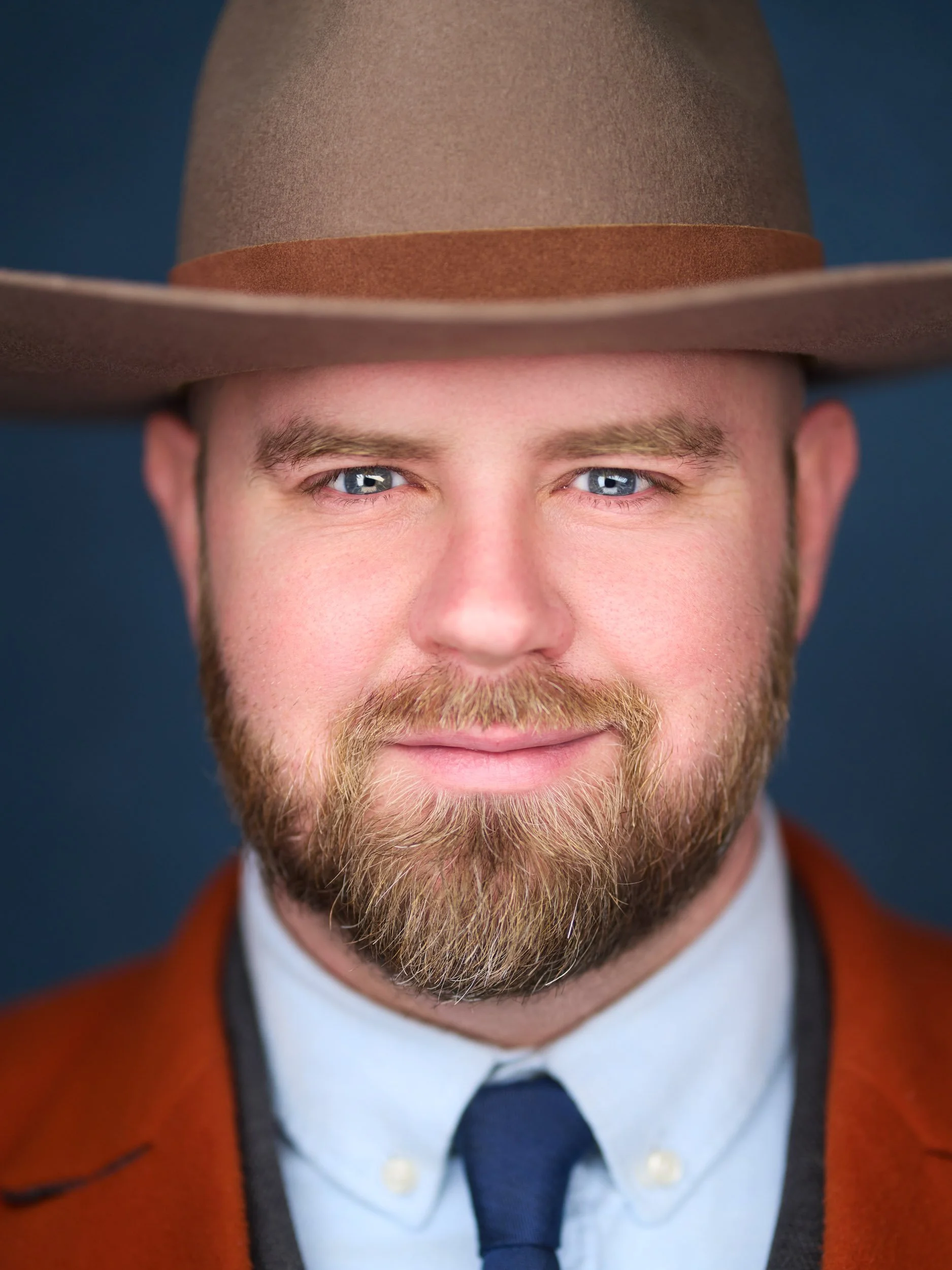 Close-up of a man wearing a brown wide-brimmed hat, white shirt, navy blue tie, and brown jacket with a dark background.