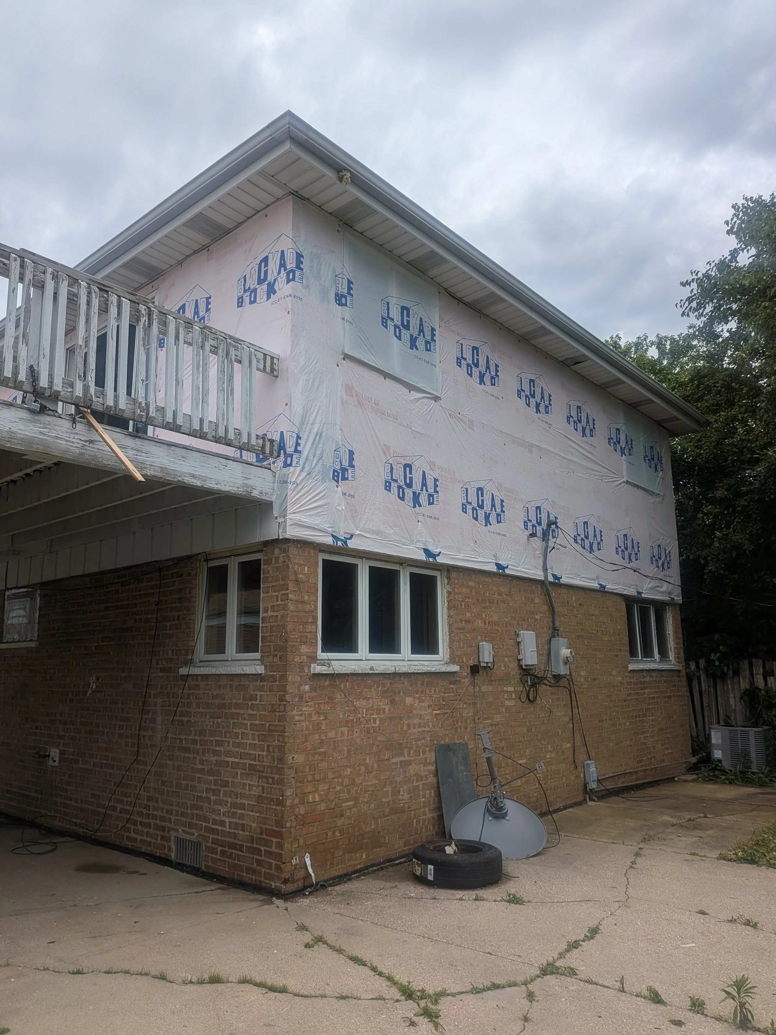 House under construction with house wrap labeled 'Lockade' on the upper story, brick exterior on the lower level, and construction tools and equipment in the yard.