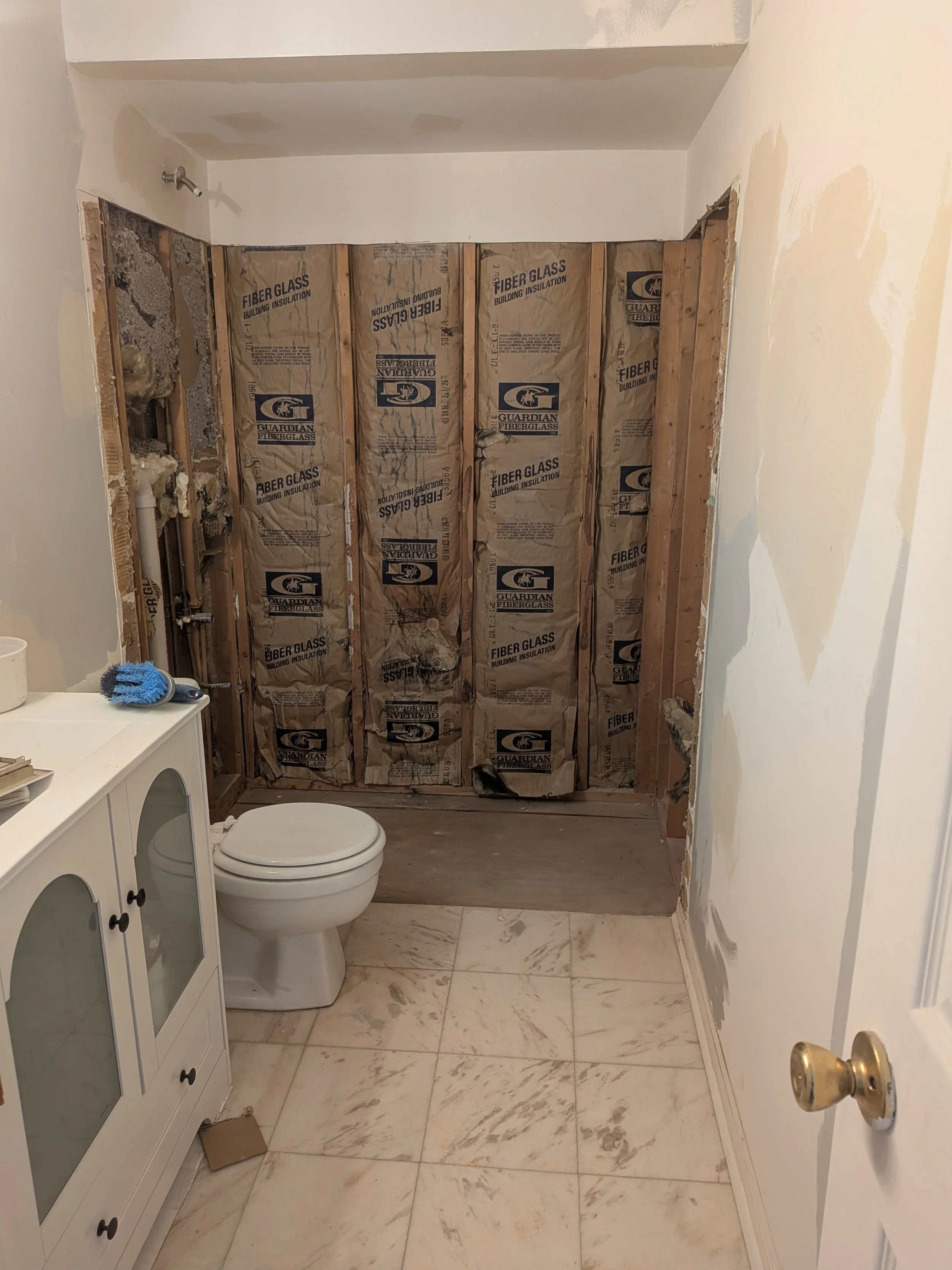 Bathroom undergoing renovation with exposed wall studs and insulation, toilet, marble tile floor, partially painted walls, and a small white cabinet with blue cleaning supplies.