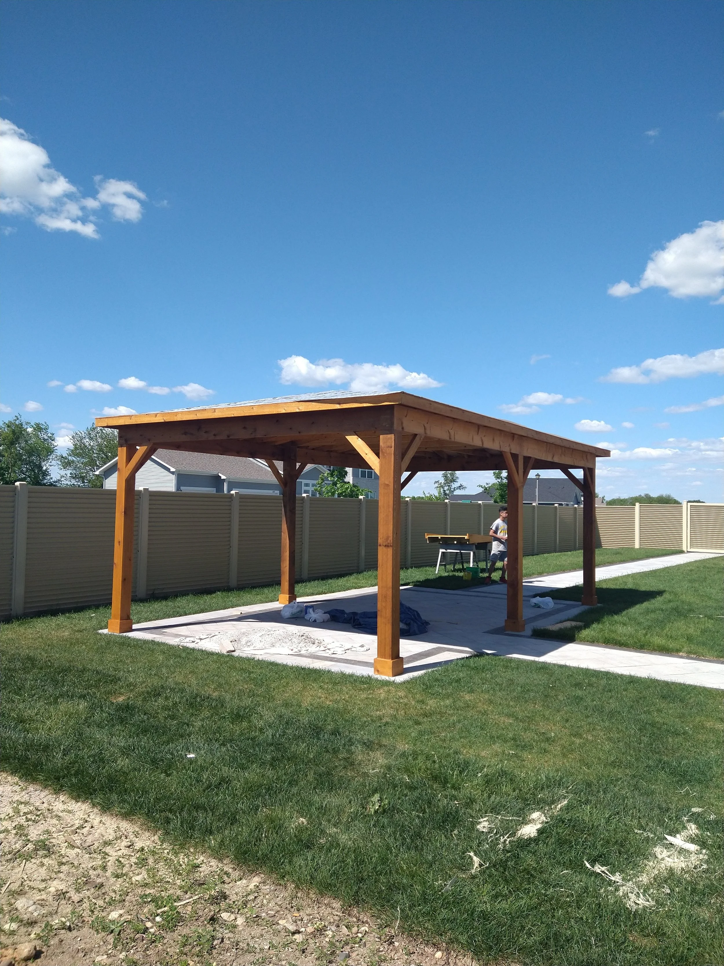 A backyard with a wooden gazebo under construction, surrounded by a beige fence, green grass, and a concrete pathway on a sunny day with blue sky and small white clouds.
