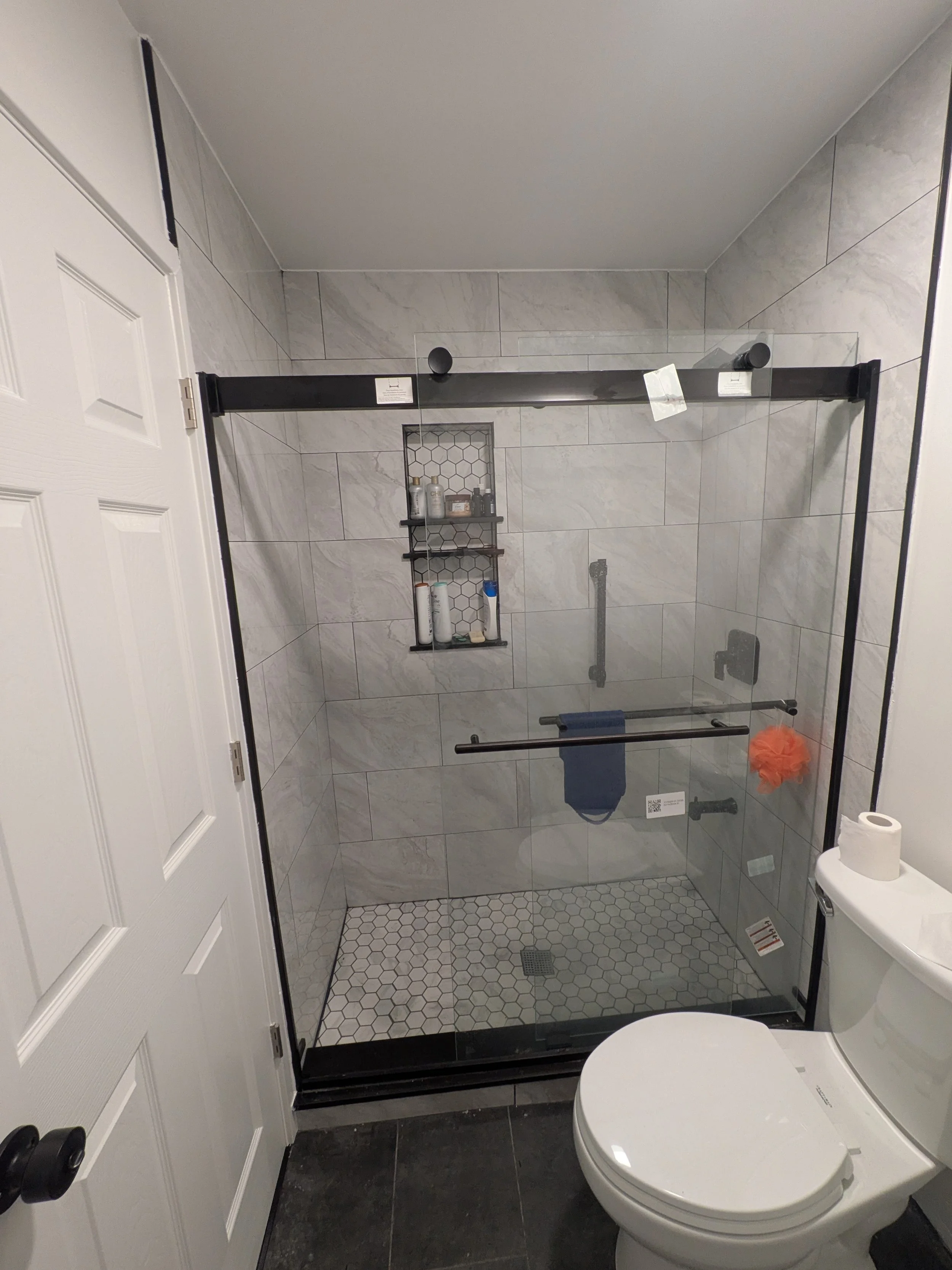 Modern bathroom with a walk-in shower enclosed by a glass door, white and gray tile walls, a built-in niche for toiletries, a gray tiled floor, a white toilet with a roll of toilet paper on top, and a closed white door.