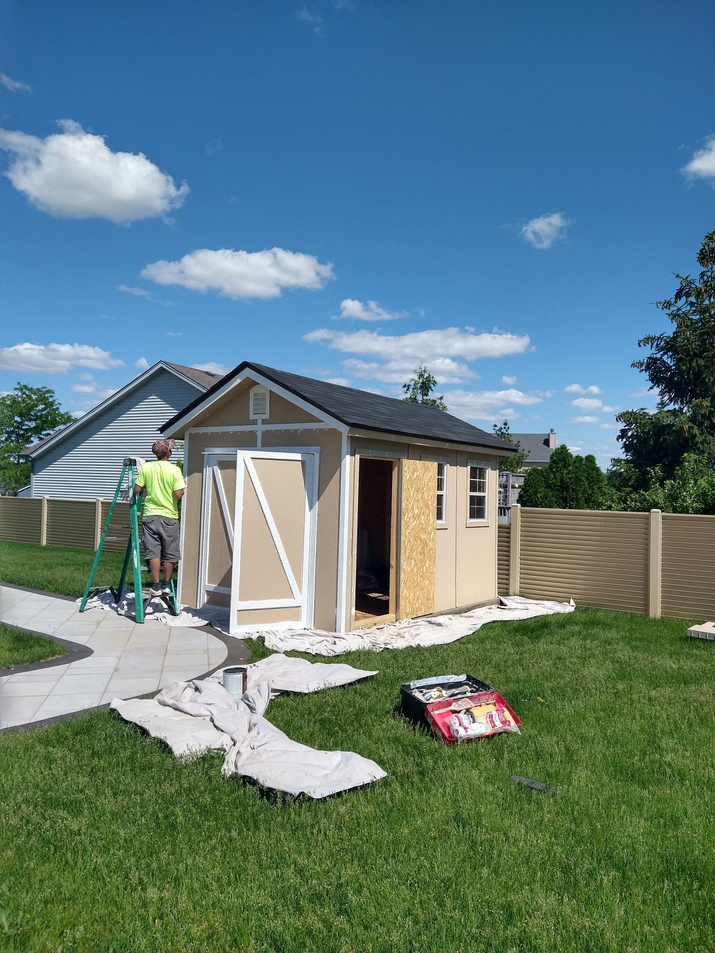 A person working on building a small shed in a backyard on a sunny day with blue sky and scattered clouds, surrounded by green grass and residential houses.