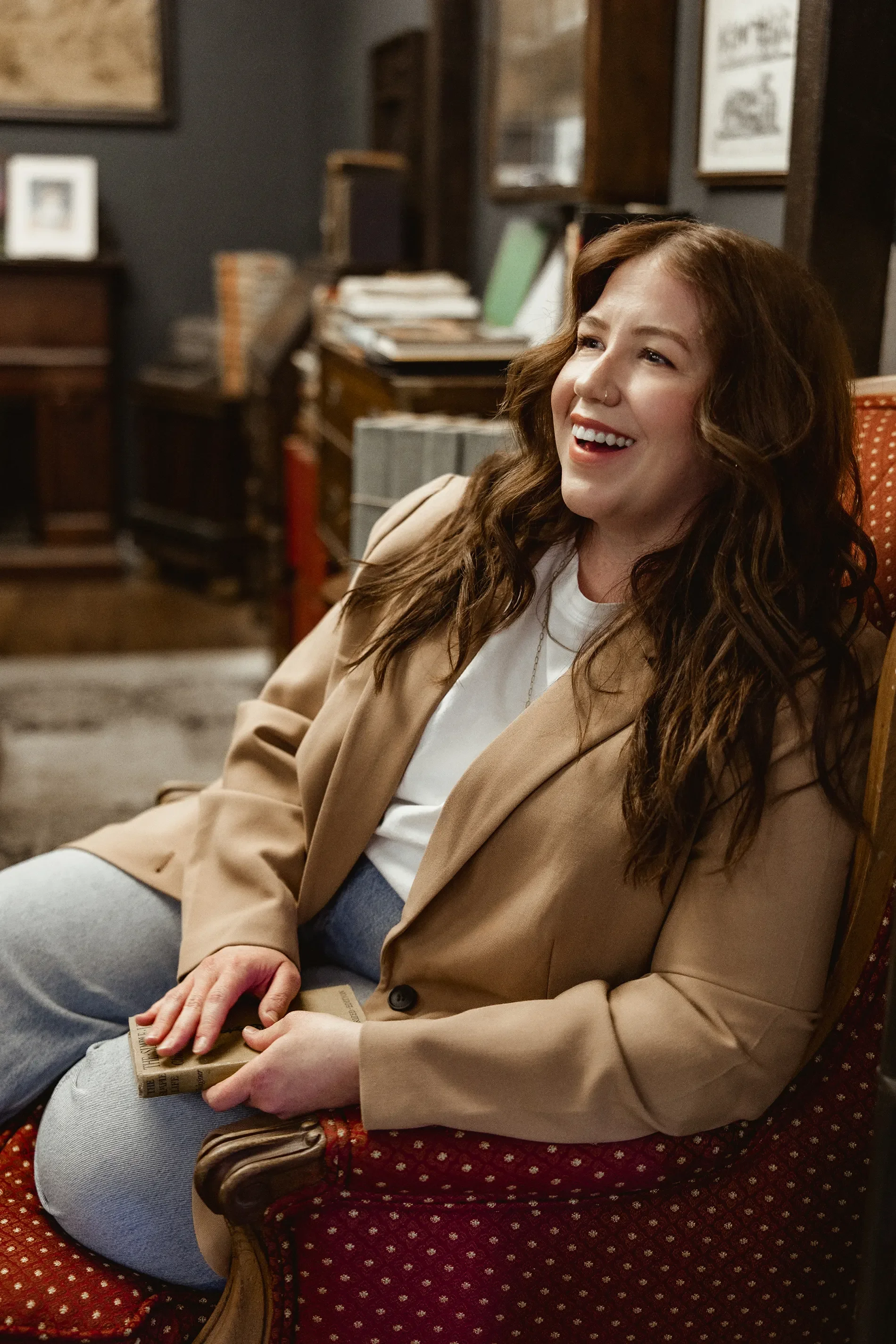 A woman with long brown hair, wearing a beige blazer, white shirt, and light blue jeans, is sitting on a vintage red patterned upholstered armchair in a cozy room with wooden shelves and books in the background. She is smiling and holding a passport or travel document.