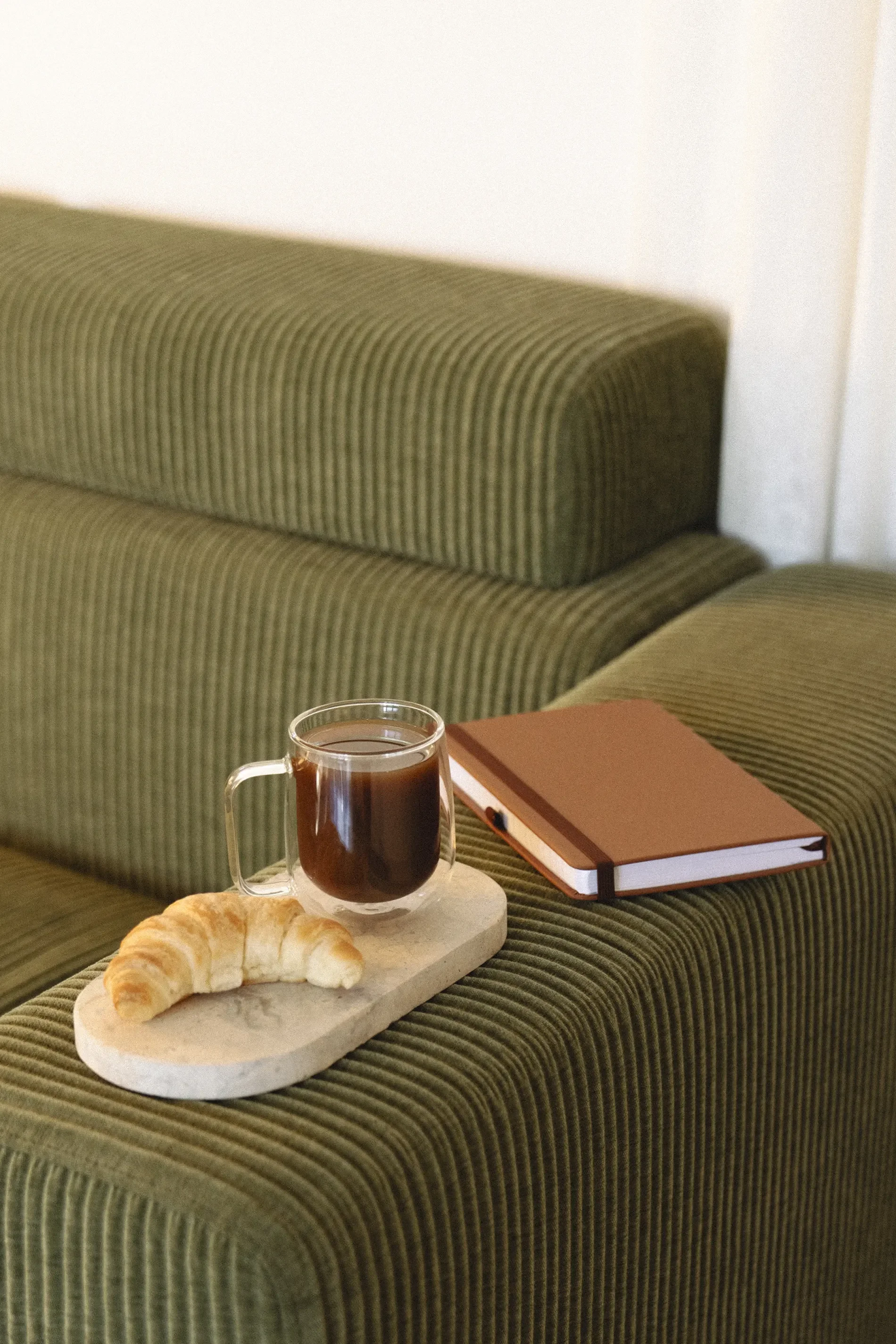 A croissant and a glass mug of coffee on a small round marble tray, with a closed pink notebook on a green-striped sofa.