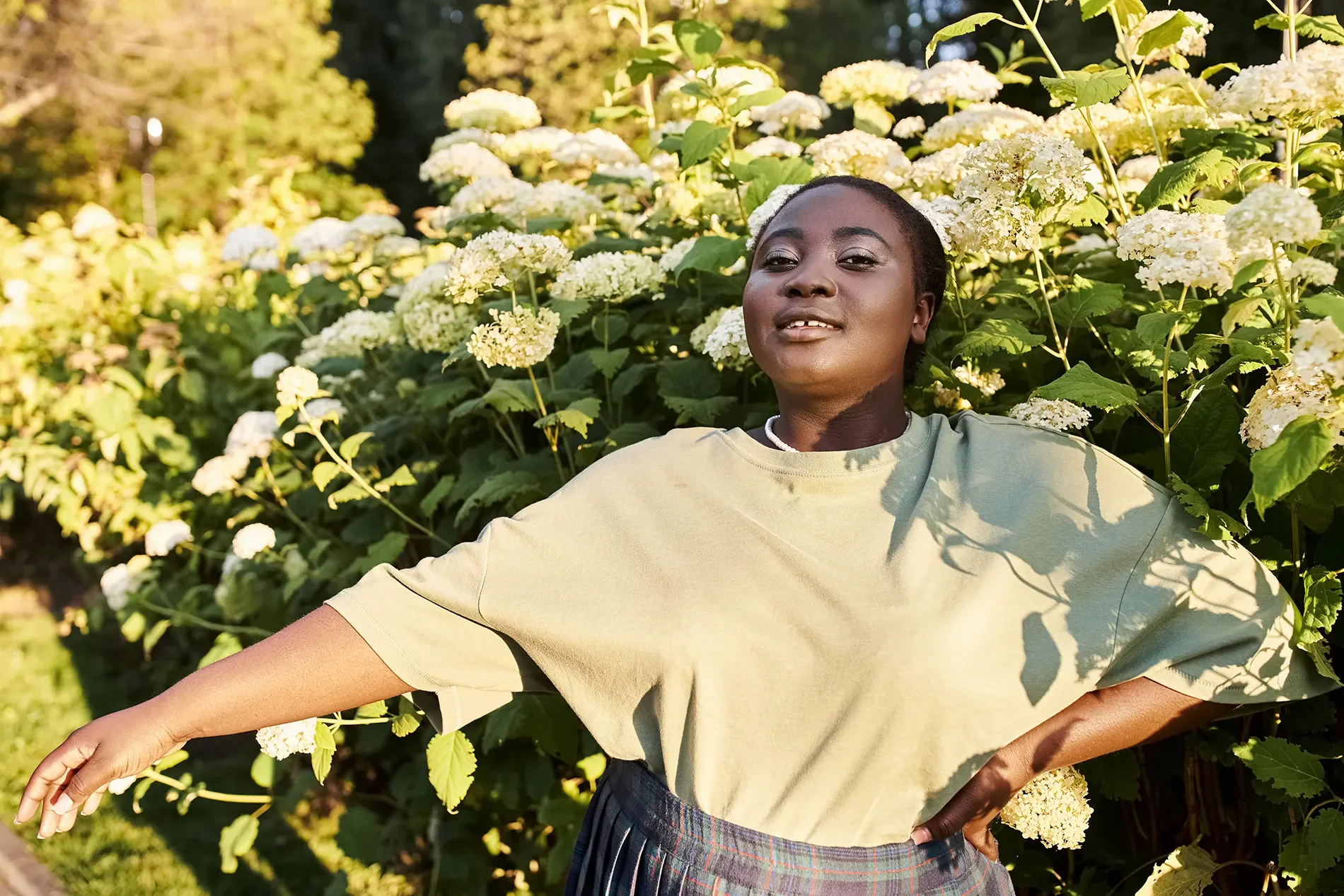 A woman with short dark hair and a dark skin tone standing among lush, white and cream-colored hydrangea flowers outdoors. She is wearing a pale yellow T-shirt and a plaid skirt, with her arms extended sideways and a confident, relaxed expression on her face.