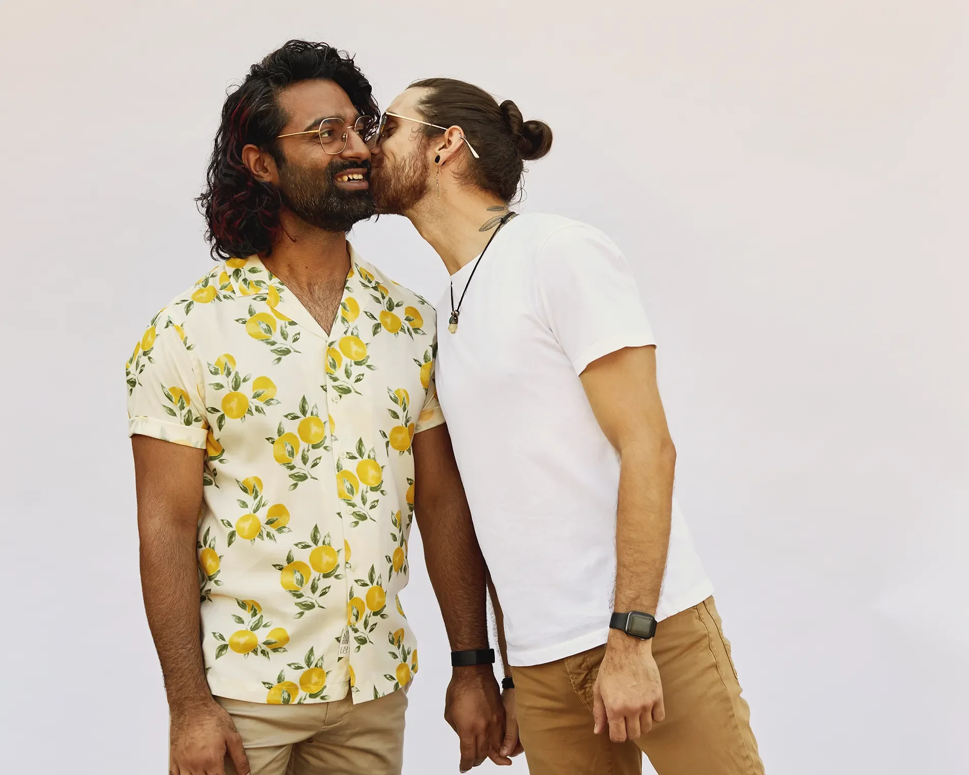 Two men stand close together, with one whispering into the other's ear. One man has long dark hair with red streaks, beard, glasses, and is wearing a white shirt with a lemon print. The other man has long hair tied back, glasses, earrings, and is wearing a white t-shirt, khaki shorts, and a smartwatch.
