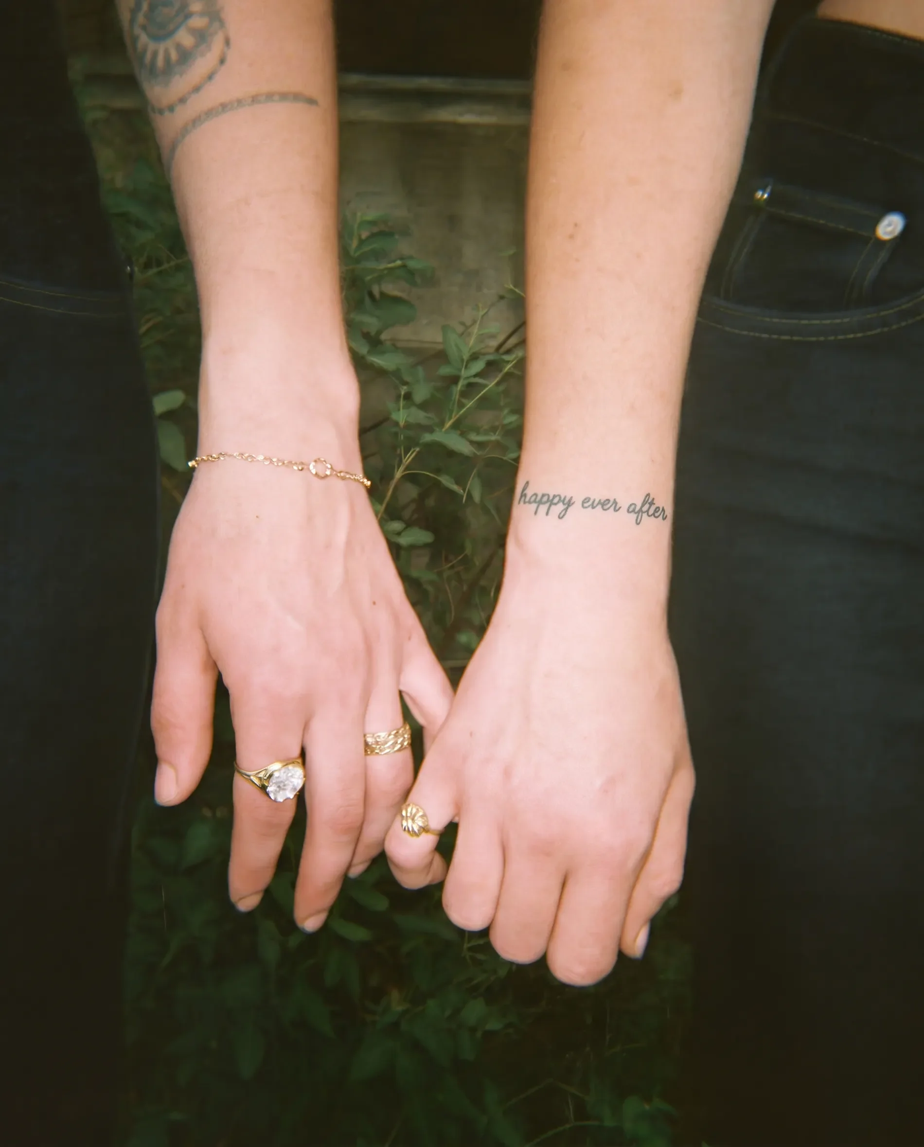 Two hands, one with multiple rings and a delicate bracelet, hold each other above green foliage. One hand has a tattoo reading 'happy ever after' on the wrist.