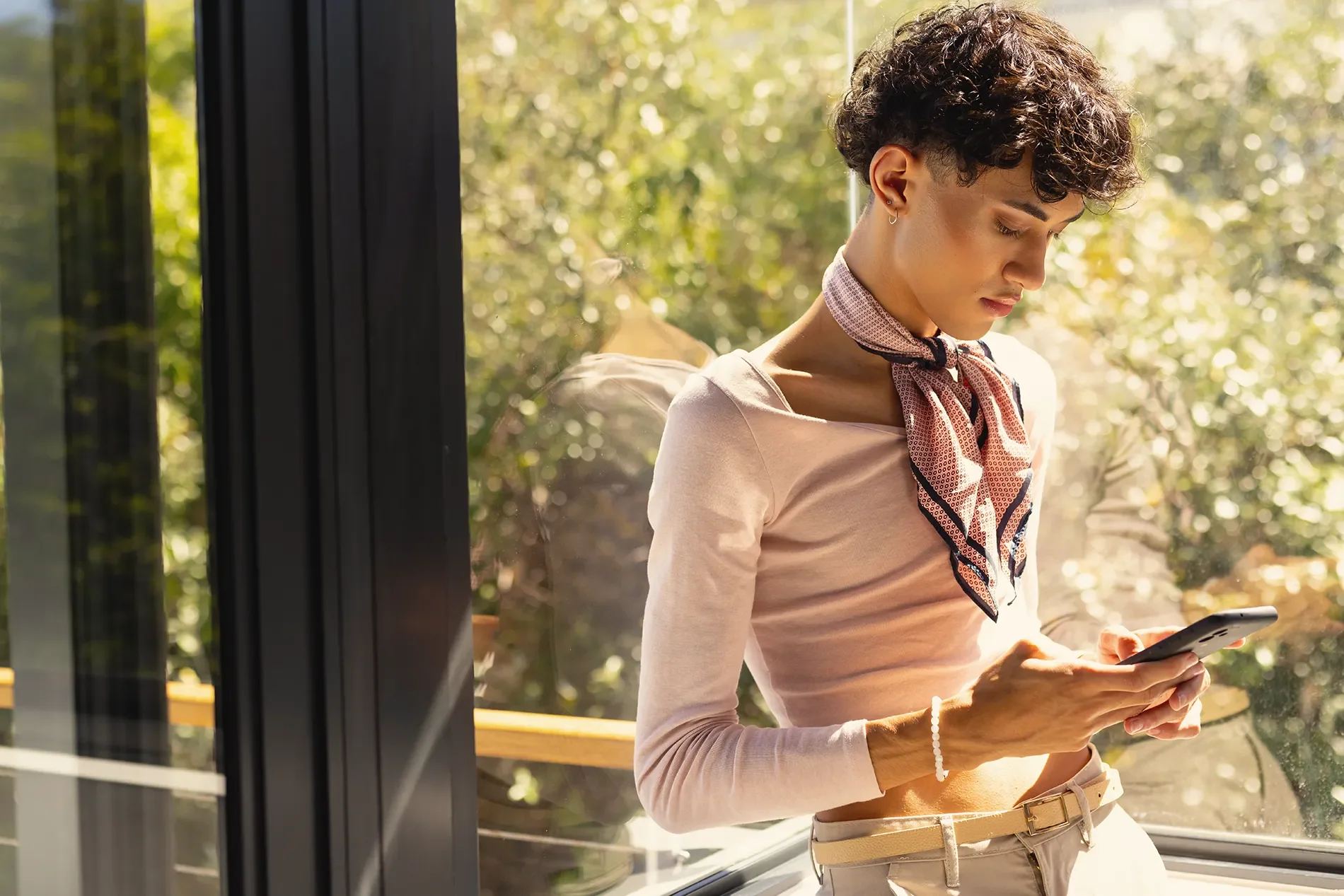 A young woman with short, curly hair and earrings, standing by a window, looking at her phone, with trees and sunlight outside.