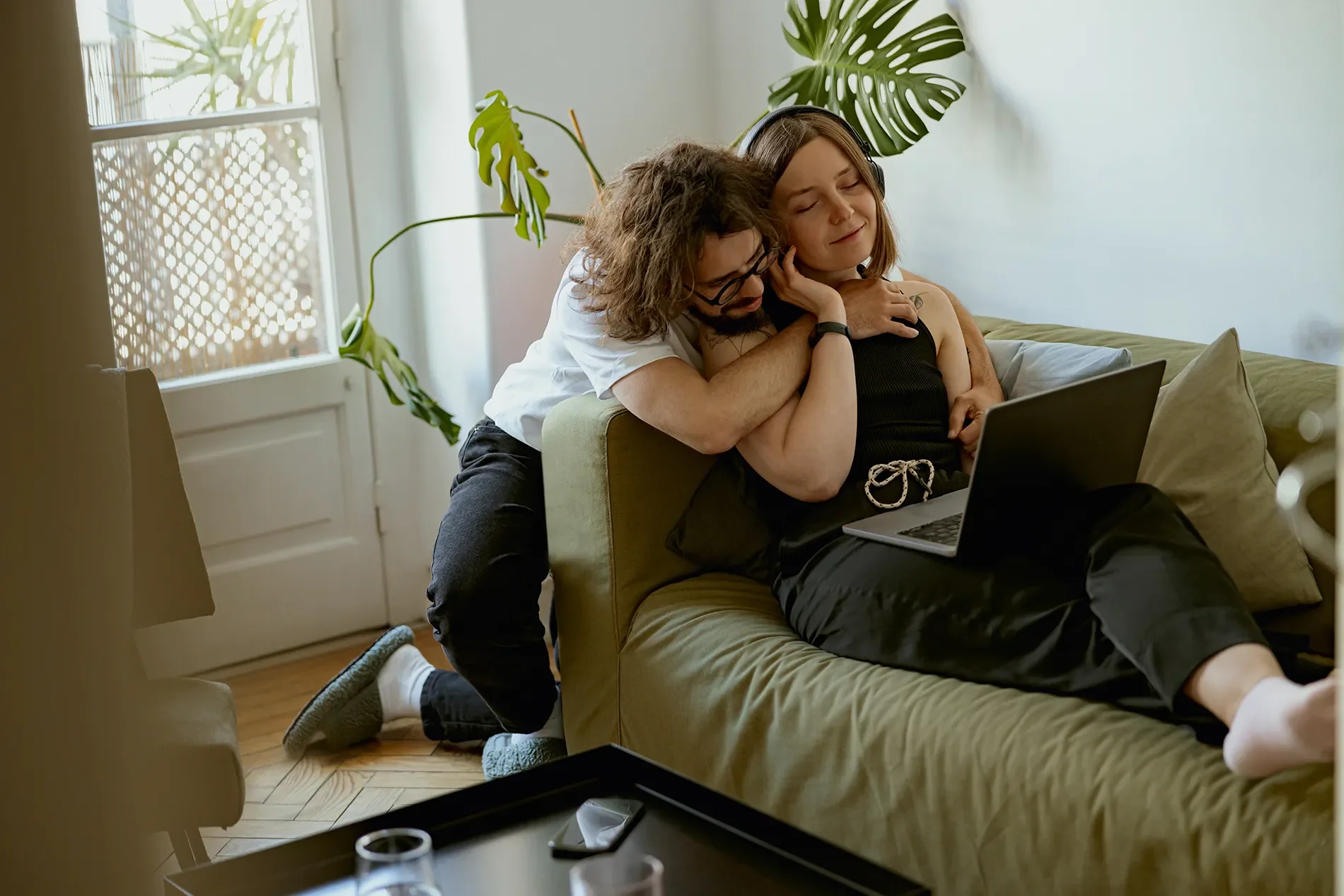 A young woman with headphones sitting on a couch, smiling with her eyes closed as a young man embraces her from behind, both looking at a laptop. The scene is indoors with a large green plant and a window in the background.