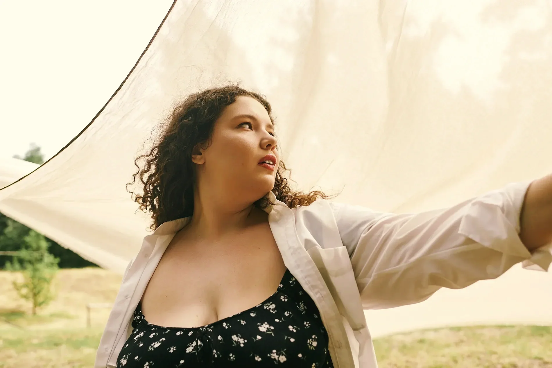 A woman with curly hair taking a selfie outdoors under a large beige umbrella.