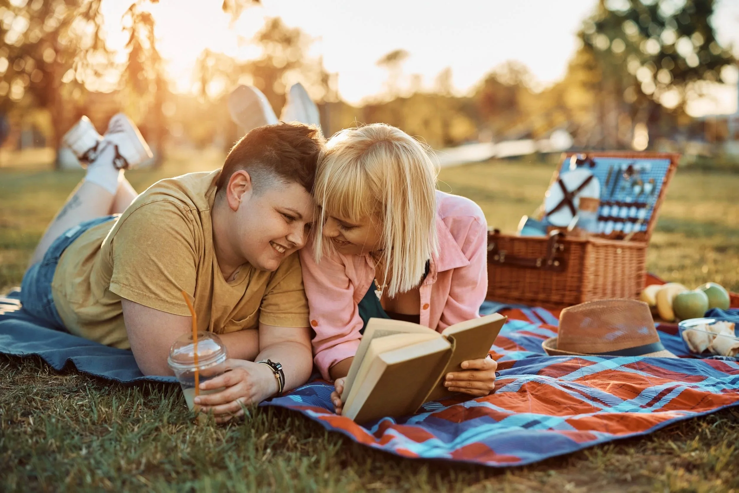 Two children, a boy and a girl, are lying on a blanket in a park during sunset, reading a book together.
