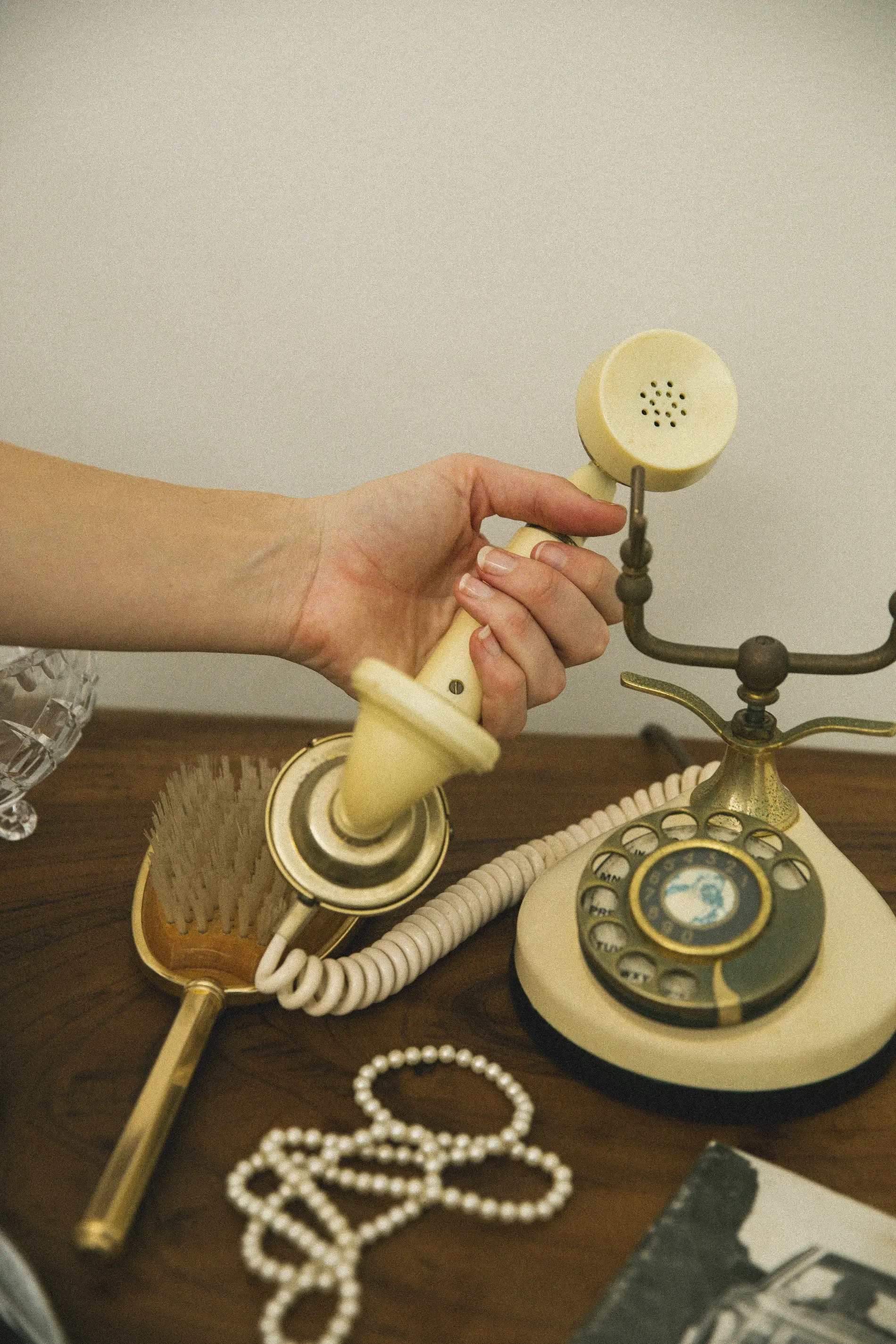 Close-up of a vintage rotary telephone, a hand holding the receiver, pearl necklaces, a glass bowl with jewelry, and other jewelry items on a wooden surface.