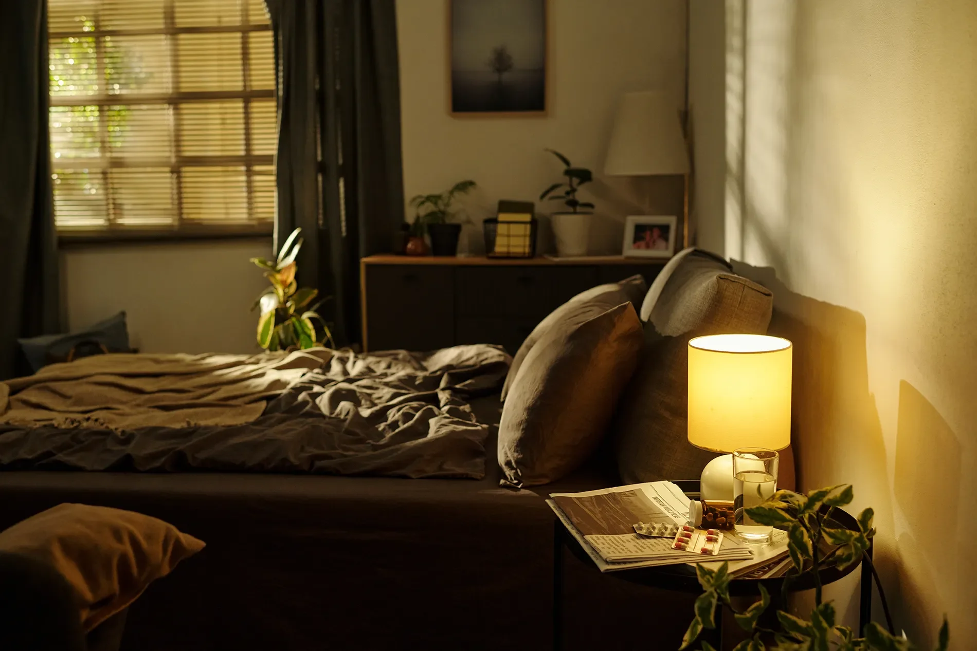 A cozy bedroom with a bed covered in dark and tan blankets, illuminated by a yellow bedside lamp. There is a nightstand with a glass, pills, and a newspaper. The wall behind the bed is painted yellow, and a small table holds a potted plant, a framed photo, and a lamp. A window with wood blinds and dark curtains is seen in the background, with sunlight filtering through.