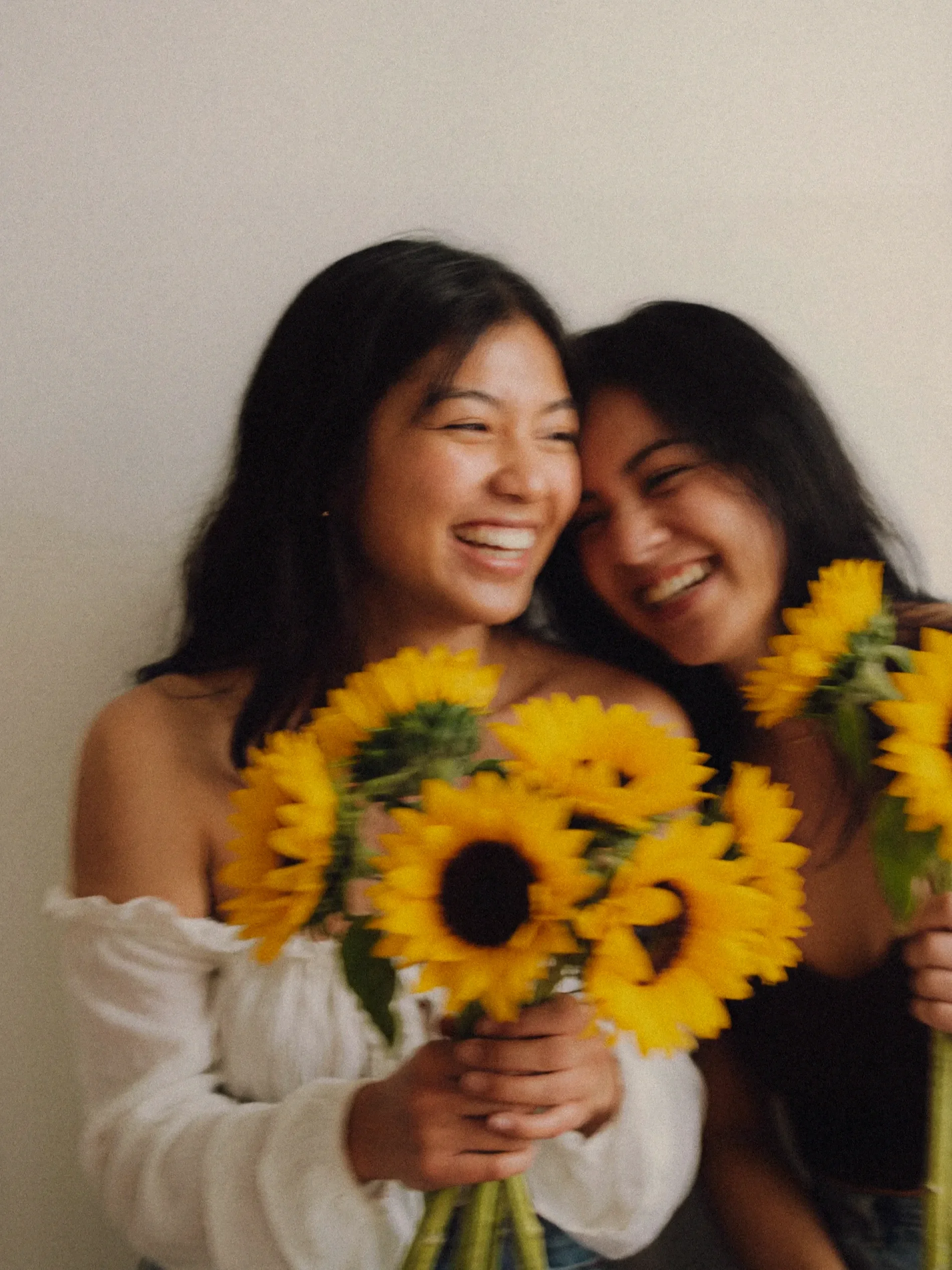 Two women smiling and hugging while holding a bouquet of yellow sunflowers against a plain background.