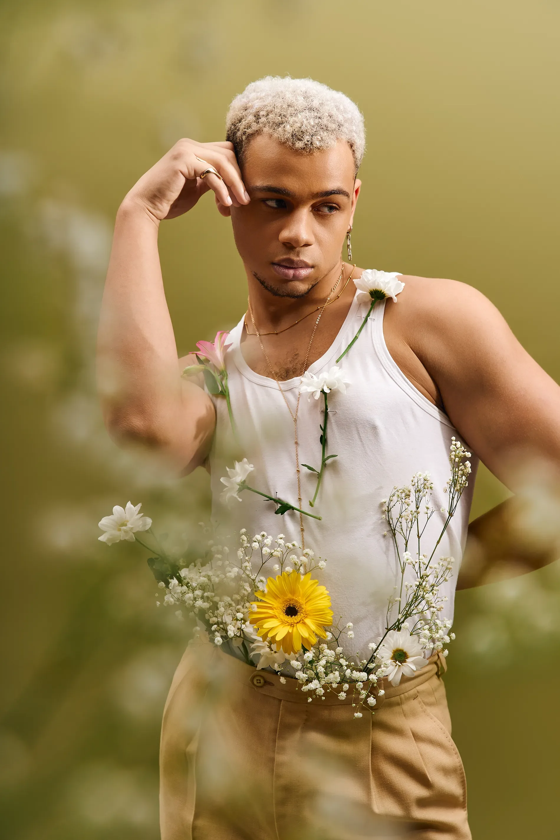 A person with blond curly hair, wearing a white tank top and beige pants, is standing in a field of flowers. They have jewelry including earrings, necklaces, and rings, and have flowers draped over their shoulder and around their waist.