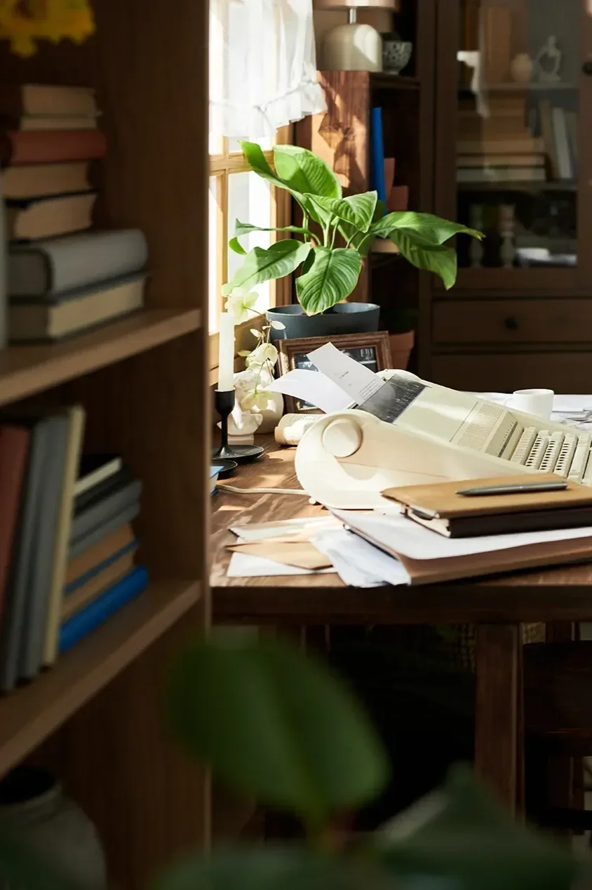 A cluttered wooden desk in a home office with a vintage typewriter, documents, and a framed photo, surrounded by bookshelves and a green potted plant in front of a window.