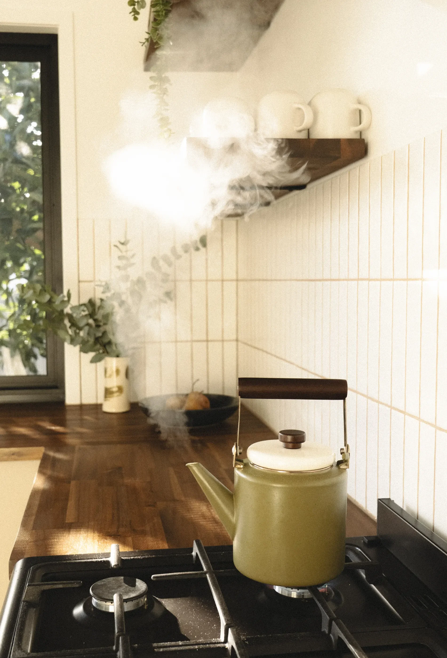 On a stove, a yellow teapot is boiling with steam coming out of the spout, in a kitchen with a tiled wall, wooden cabinets, a window, and various kitchen items.