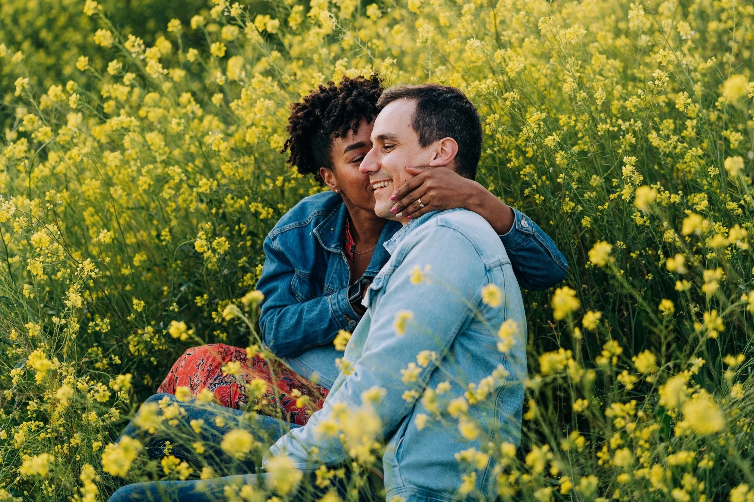 A couple sitting in a yellow flower field, sharing a kiss and smiling.