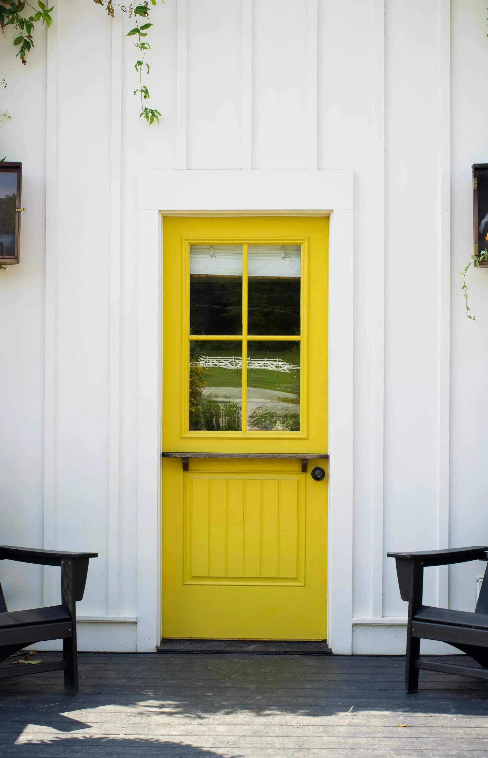 A white exterior wall with vertical siding and a bright yellow door with window panes, flanked by two black outdoor chairs on a wooden porch.